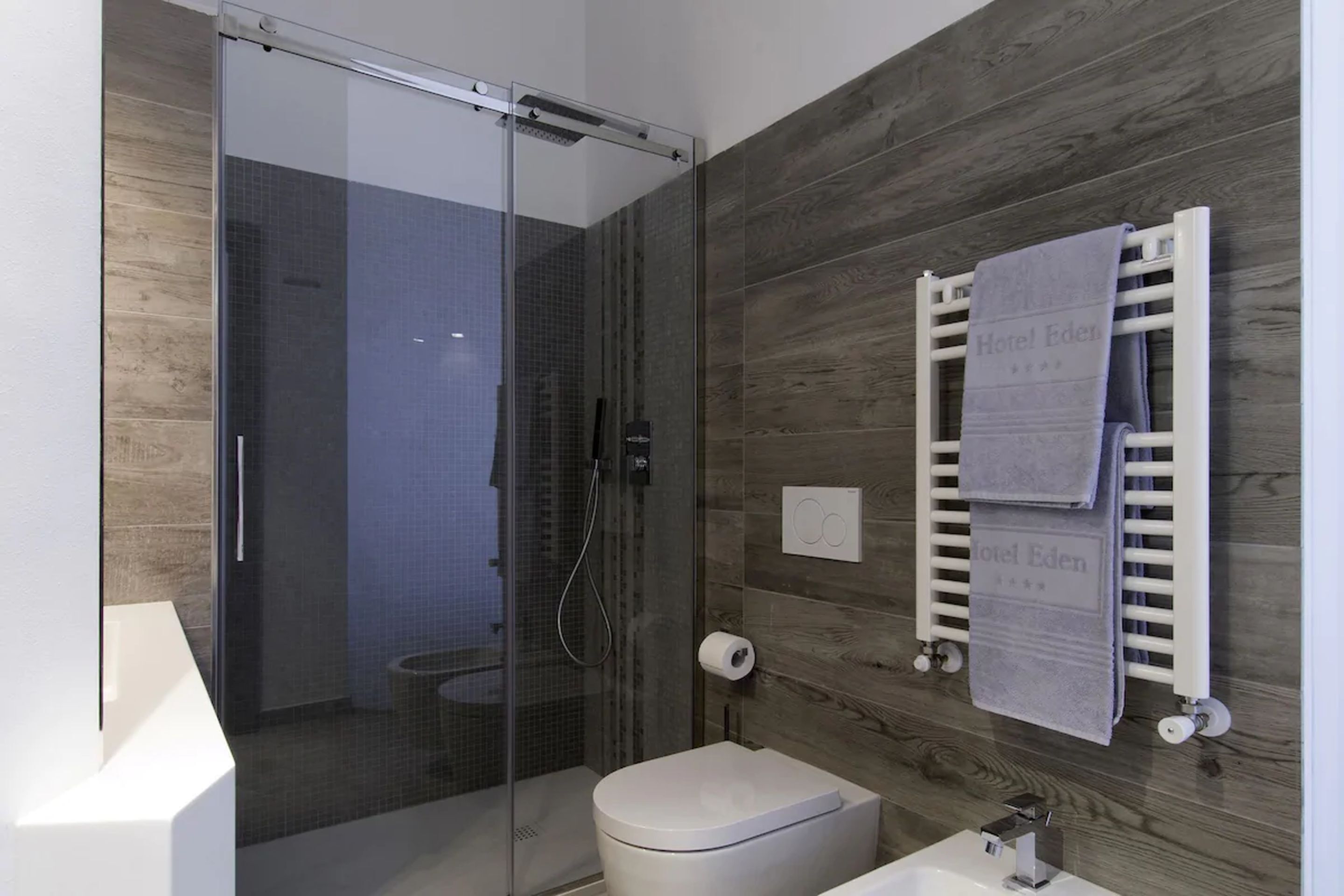 A modern bathroom with a glass-enclosed shower, a white toilet, and a towel rack with a towel labeled "Hotel Eden."