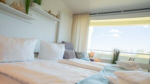 A hotel room with a large bed, pillows, a bedside table with towels, a window with a view, and a shelf with decorative items. - Apartment 254 Little time-out (ID 139) (Sankt Peter-Ording)