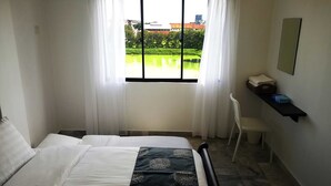 Et hotelværelse med en seng, en stol, et lille bord og et vindue med udsigt over et grønt område og bygninger.