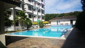 En swimmingpool med flisebelagt dæk, omgivet af en bygning med flere etager, altaner og en lille bygning med tag.