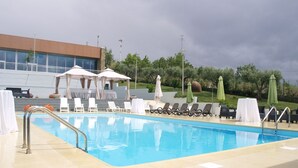 Una piscina con sillones de descanso y sombrillas, un edificio al fondo y un cielo nublado.