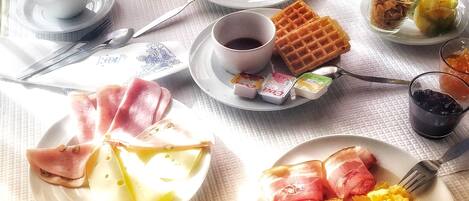 Une table dressée pour le petit-déjeuner, avec différents plats, notamment des toasts, des œufs, du jambon, du fromage et des fruits.