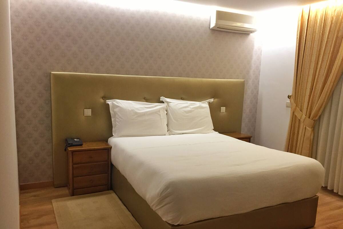 Una cama bien hecha con cabecera, mesita de noche y pequeña cómodita.