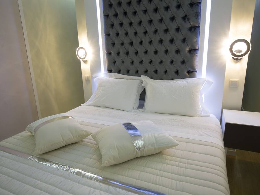 Een netjes opgemaakt bed met witte lakens en kussens, een gestoffeerd hoofdbord en bedlampjes.