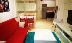 Un ambiente compatto con un divano rosso, un tavolino da caffè bianco e un'area pranzo con sedie rosse. Si intravede in secondo piano una cucina con un frigorifero e un televisore.