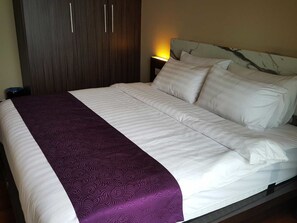 Ein ordentlich bezogenes Bett mit weißer und violetter Tagesdecke, ein Holzschrank und ein Nachttisch mit Lampe.