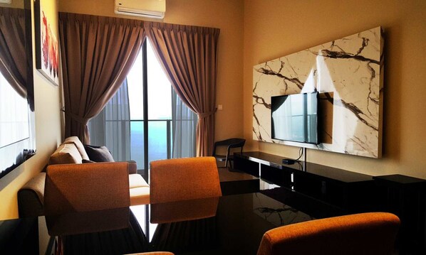 Quarto de hotel com uma janela grande, uma TV de tela plana na parede e uma área de estar com duas poltronas laranjas e um sofá.