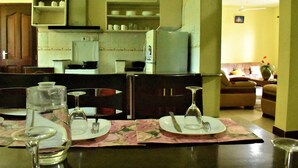 A dining area with a table set for two, a kitchen in the background, and a living room with a sofa and a vase of flowers. - Suswa Six Apartment (Mombasa)
