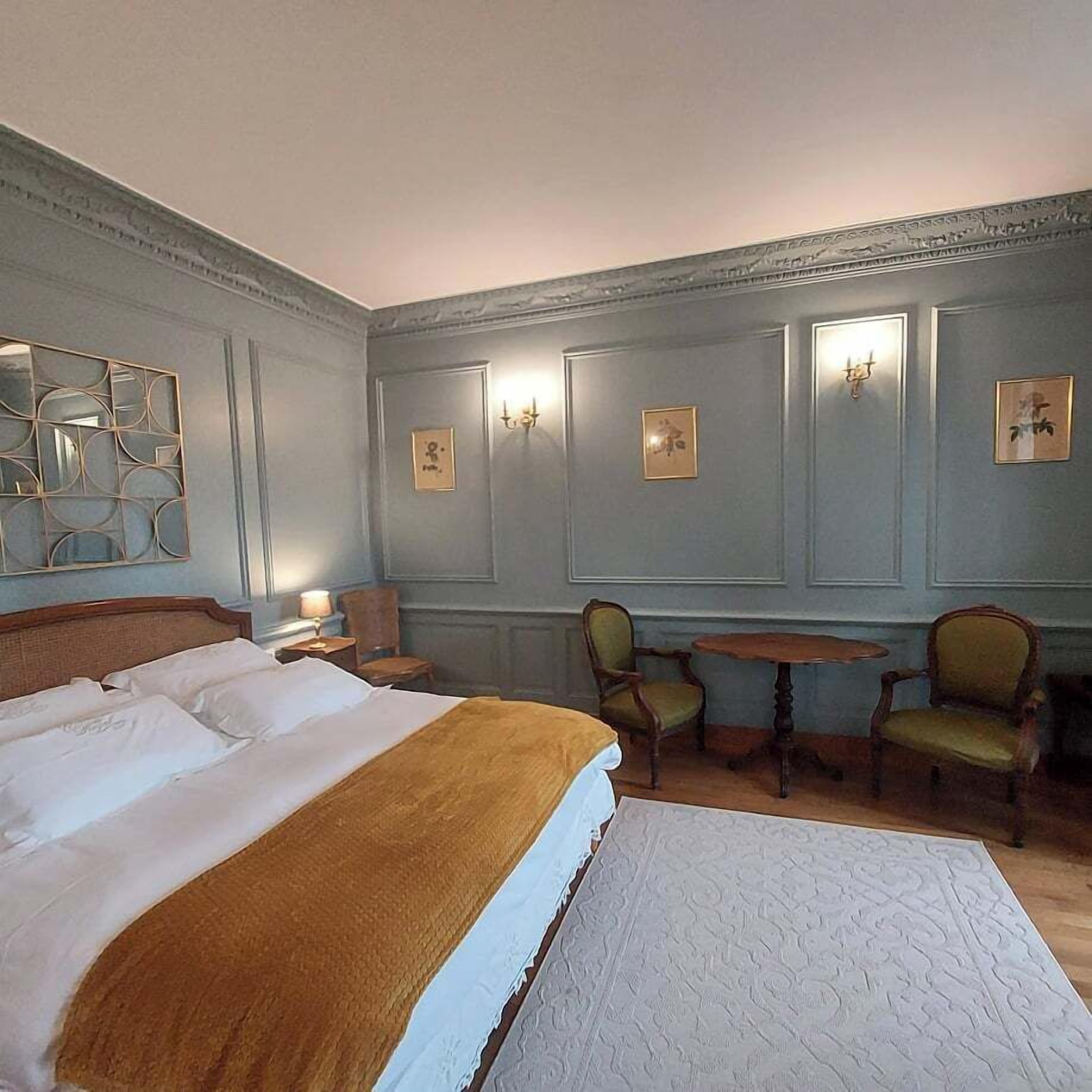 A hotel room with a large bed, two chairs, a small table, and wall-mounted sconces.