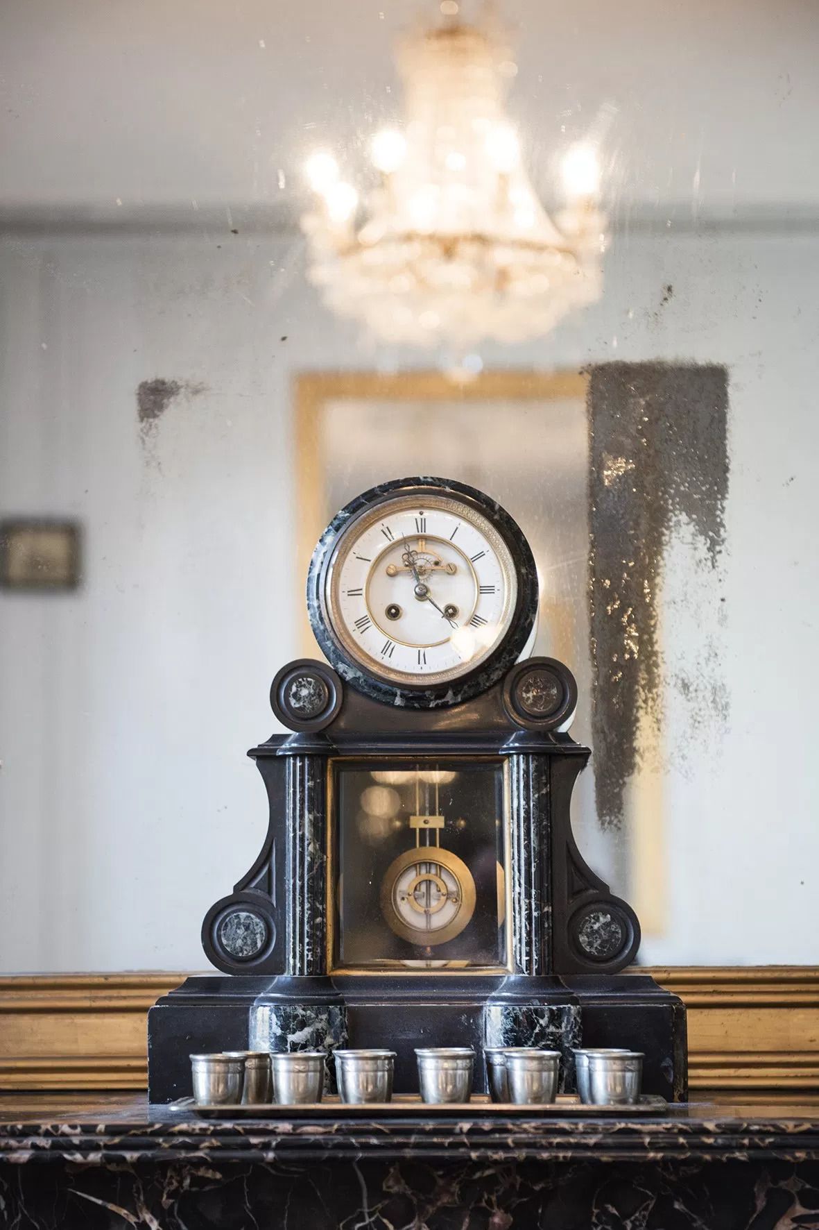 A vintage clock with Roman numerals on a marble base, surrounded by metal cups on a marble countertop.