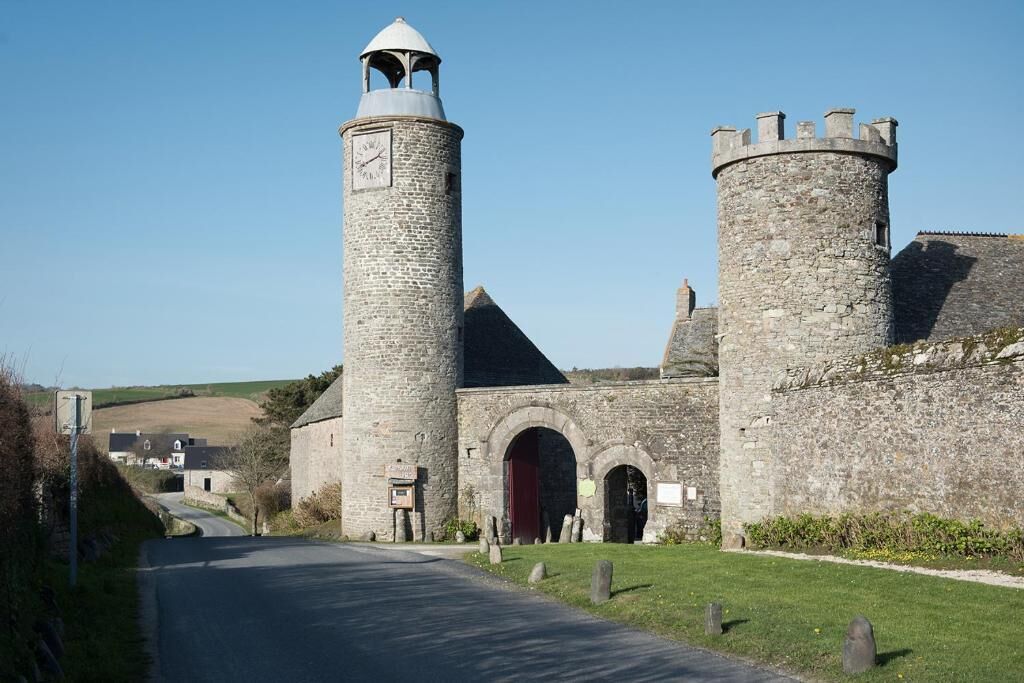 A historic stone building with a clock tower and two round towers, featuring an arched entrance and a paved pathway leading to it.