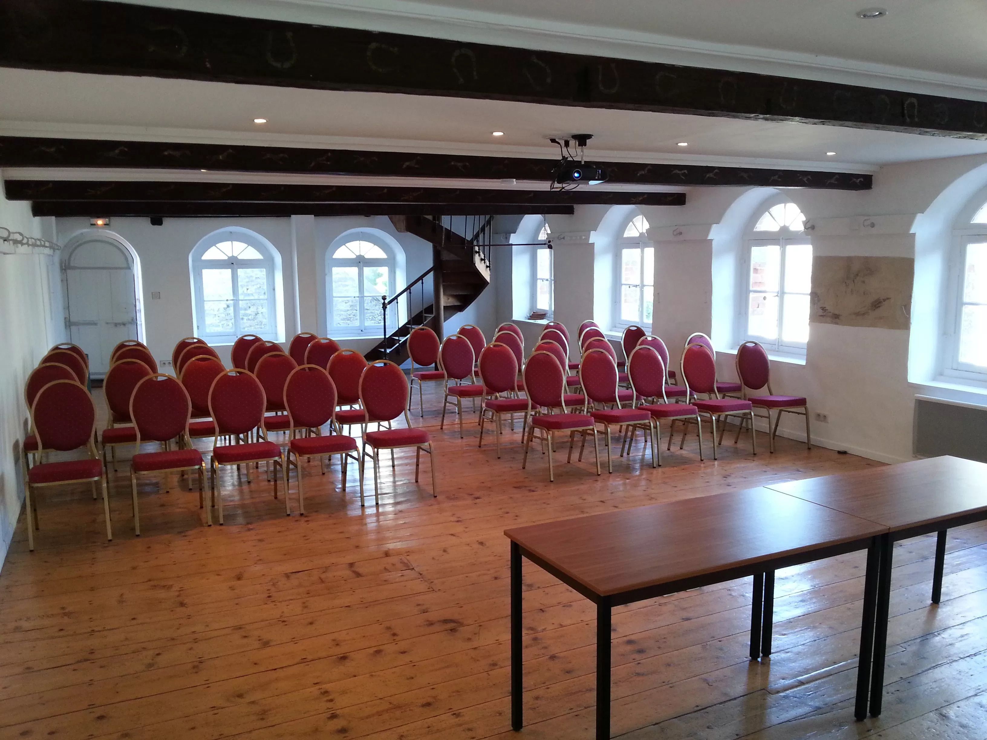 Une salle de conférence avec des rangées de chaises rouges, une table en bois et un escalier menant à un niveau supérieur.