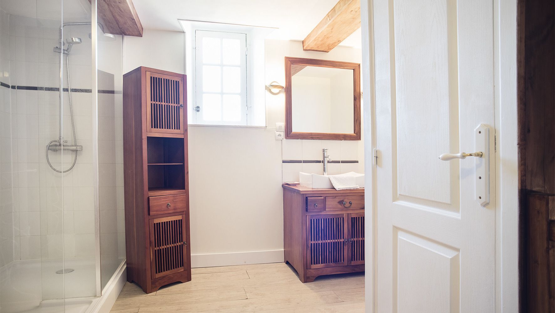 A bathroom with a glass shower enclosure, a wooden cabinet with drawers, a sink with a mirror, and a window above the sink.