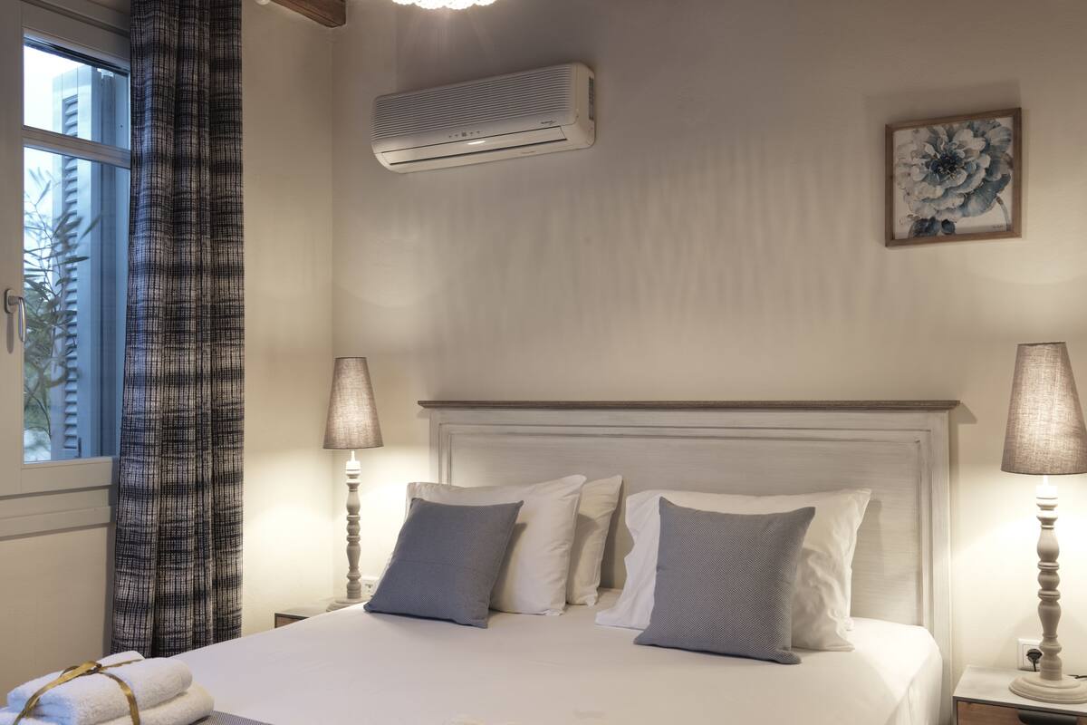 A hotel room with a bed, two bedside lamps, a window with curtains, an air conditioning unit, and a framed picture on the wall.