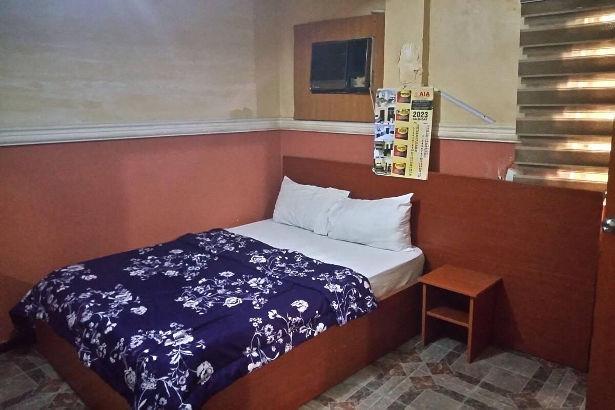 A single bed with a floral bedspread, a wooden headboard, a small wooden nightstand, a wall calendar, and a window with blinds.