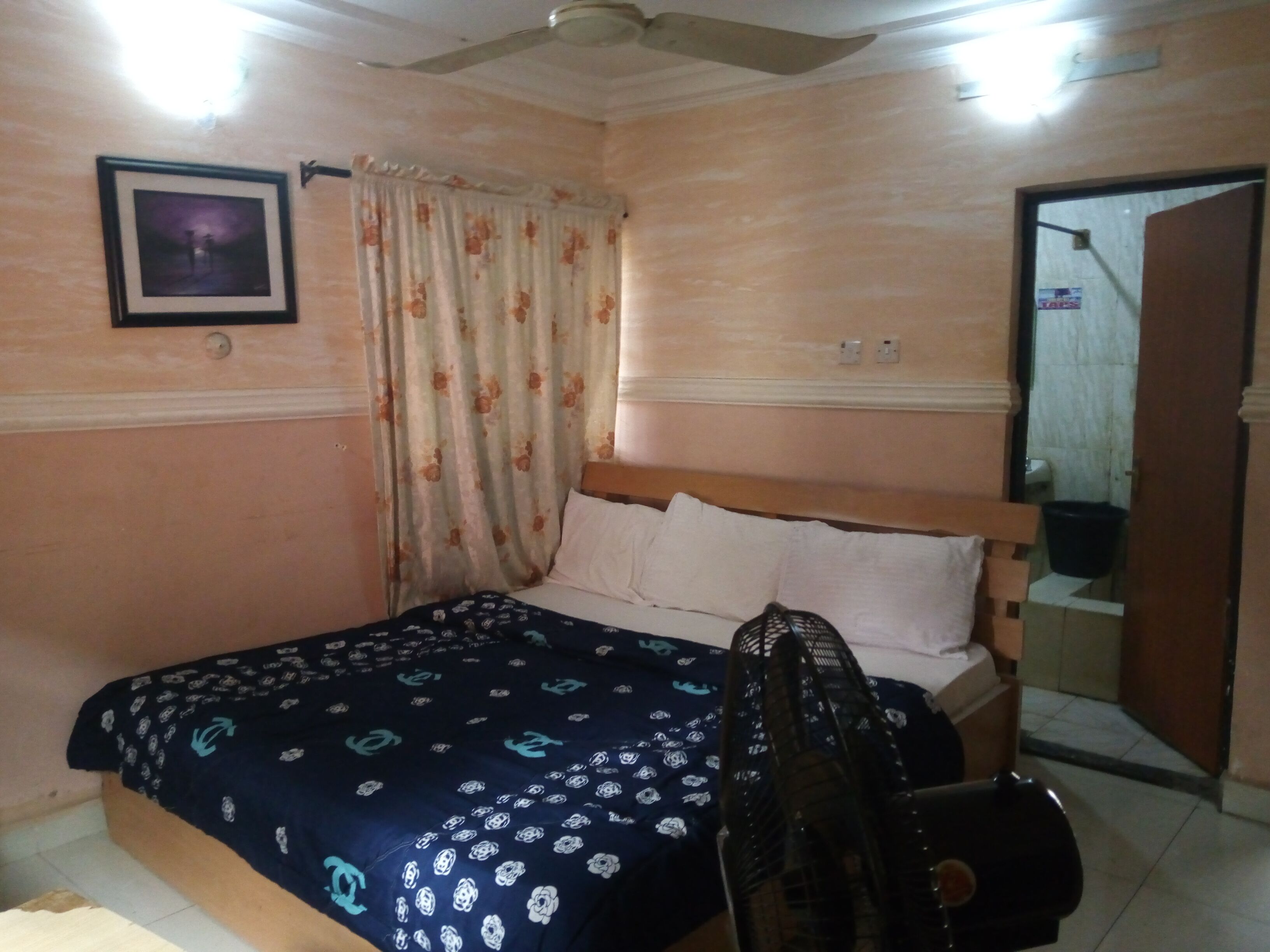 Un dormitorio con cama, ventilador de techo, ventana con cortinas florales, un cuadro en la pared y una puerta que da a otra habitación.