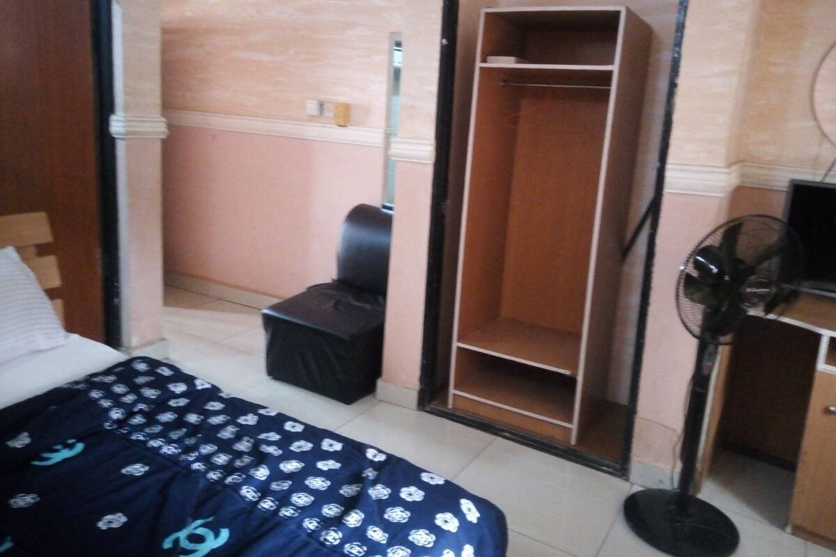 A bedroom with a bed, a wardrobe, a desk with a chair, and a standing fan.