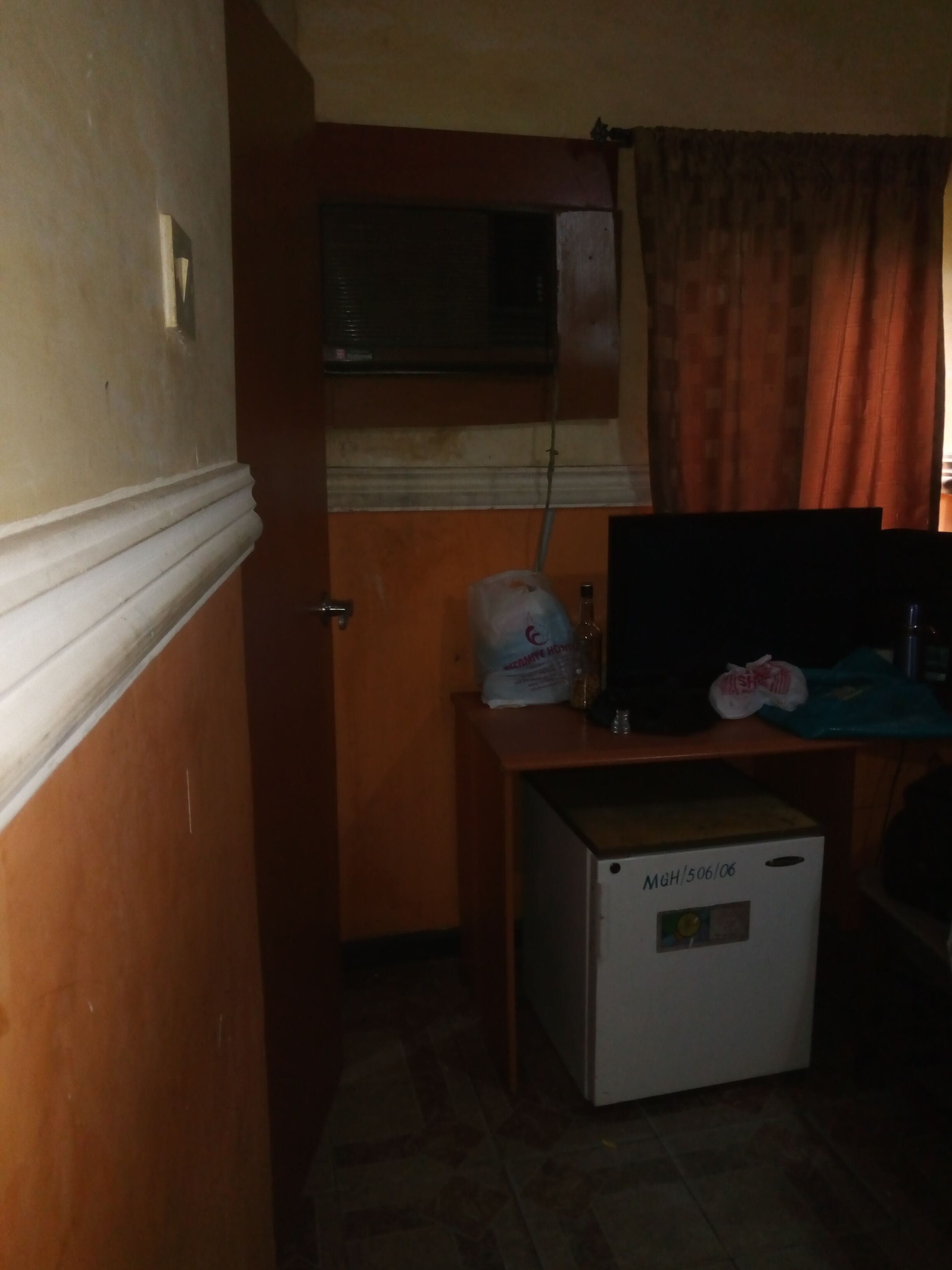 A compact room with a small refrigerator, a wooden desk with a computer monitor, a window with curtains, and a door with a light switch.