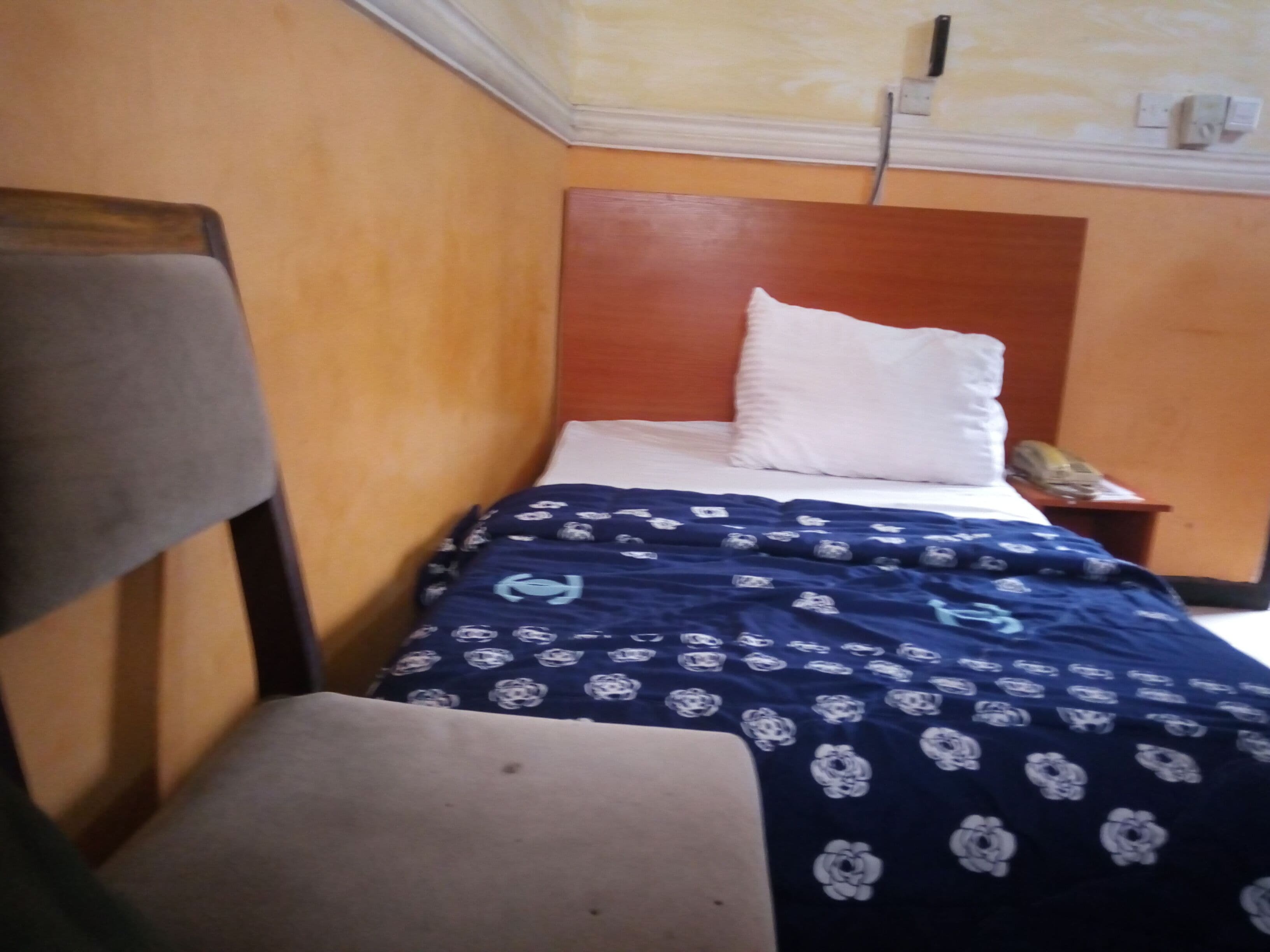 Ein kleines Zimmer mit einem Einzelbett, einem Holzkopfstück, einem weißen Kopfkissen und einer blumigen, blauen Tagesdecke.