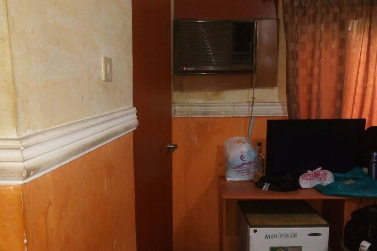 A compact room with a bed, a small desk with a computer, a refrigerator, and a window with curtains.