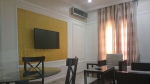 Ein Wohnzimmer mit Flachbildfernseher an einer gelben Wand, einem Glastisch mit Stühlen, einem Sofa und einem Couchtisch.