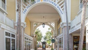 The entrance of The Village of Baytowne Wharf with a covered walkway, palm trees, and a coffee shop visible.