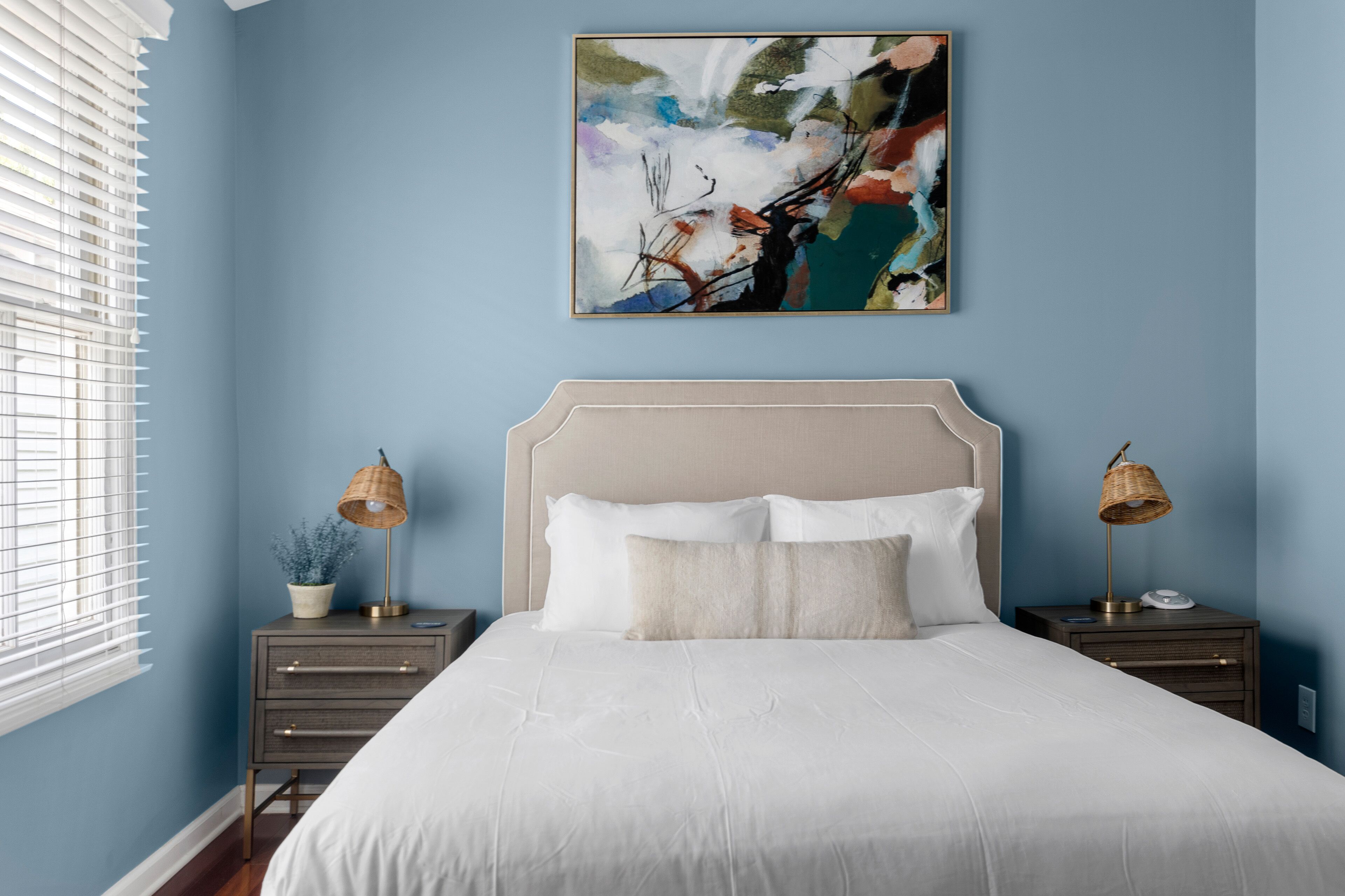 Een slaapkamer met een bed, twee nachtkastjes, een lamp, een schilderij en een raam met zonwering.
