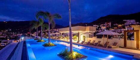 A nighttime pool area with lounge chairs and palm trees, surrounded by buildings and a mountainous backdrop.