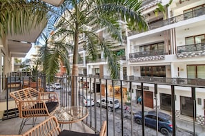 A balcony with wicker furniture, a palm tree, and a view of a street with cars and buildings.