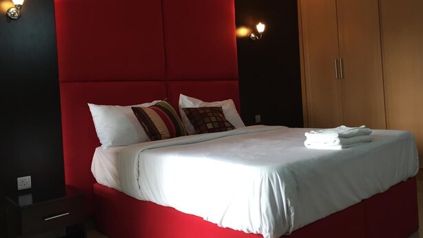 Una cama con ropa de cama blanca, cabecera roja y varias almohadas. Se observa un armario de madera y una mesita de noche con una lámpara.