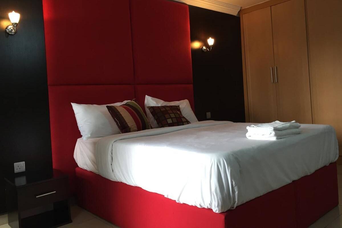 A bed with white bedding, red headboard, and multiple pillows. There is a wooden wardrobe and a nightstand with a lamp.