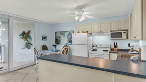 A modern kitchen with a white refrigerator, microwave, stove, and sink. There is a ceiling fan, a dining area with chairs, and a large window with a decorative bird mural. - 1 Bedroom / New Furniture / Direct beach access / Across from J.W. Marriott! (Marco Island)