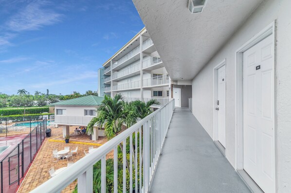 A hotel balcony with a pool, white railings, and a view of a building with multiple floors.