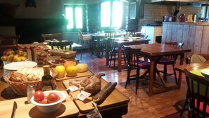 A dining area with multiple wooden tables and chairs, a kitchen in the background, and various fruits and vegetables on the tables. - Hotel Cruells (Planoles)