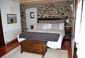 A bedroom with a stone wall, a bed with white bedding, a wooden bench, and a nightstand with a lamp.