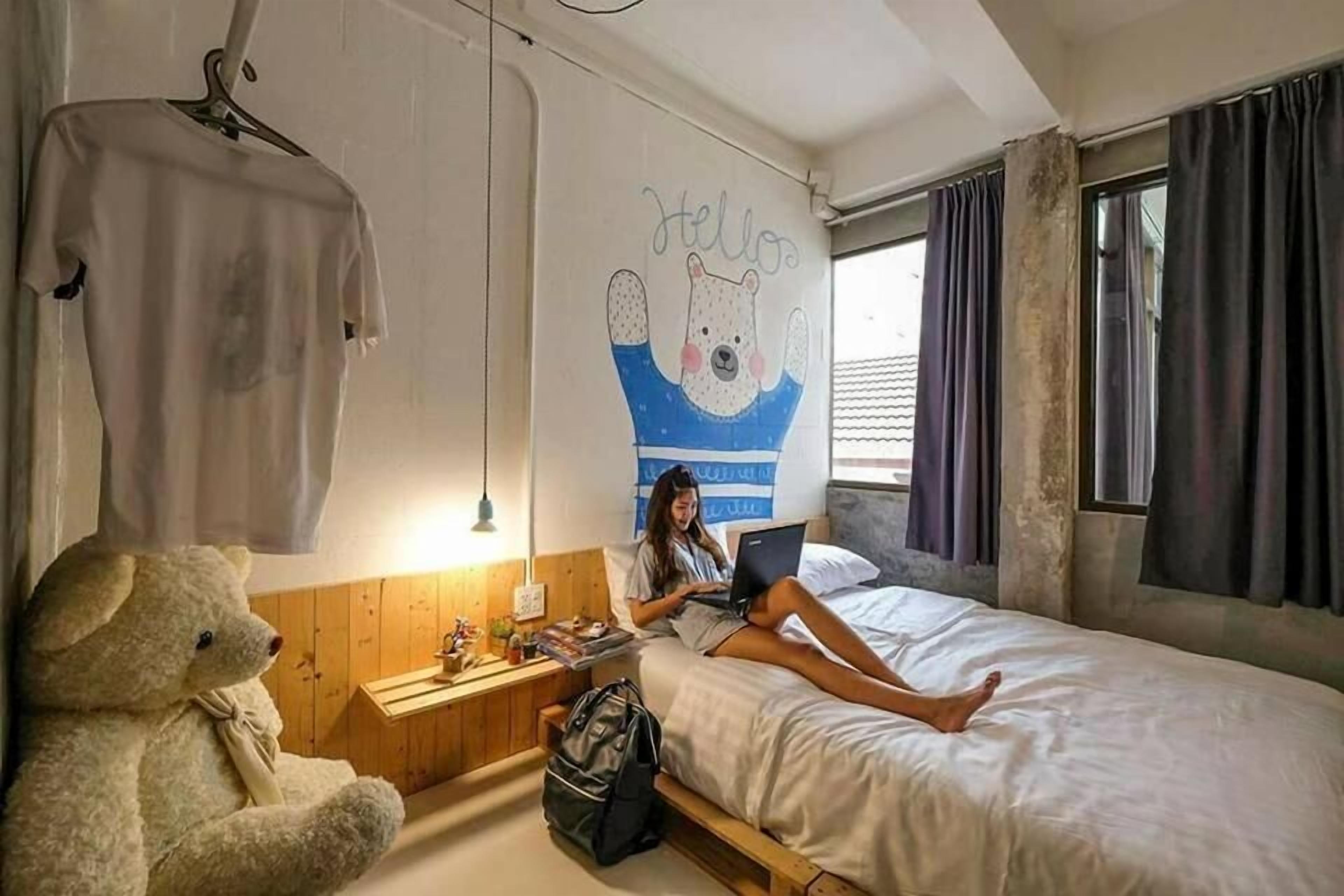 A bedroom with a bed, a teddy bear, a laptop, and a wall with a bear illustration.