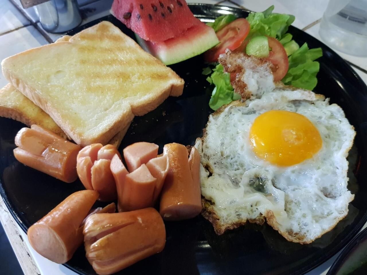 A plate with toast, sausages, a fried egg, and a slice of watermelon.