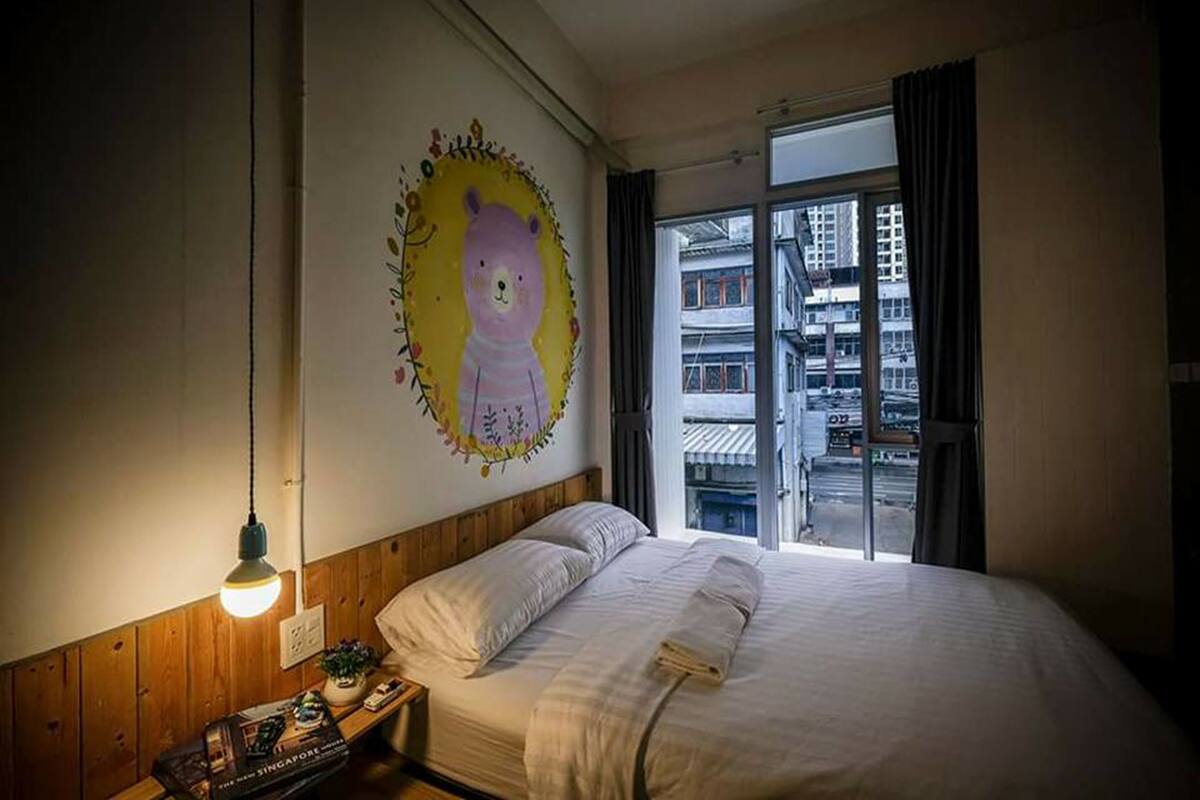 A bedroom with a bed, a nightstand, a wall painting, and a window with a view of buildings.
