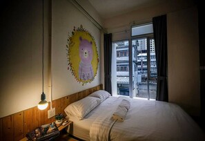 A bedroom with a bed, a nightstand, a wall painting, and a window with a view of buildings. - Pastel House - Adults Only - Hostel (Bangkok)