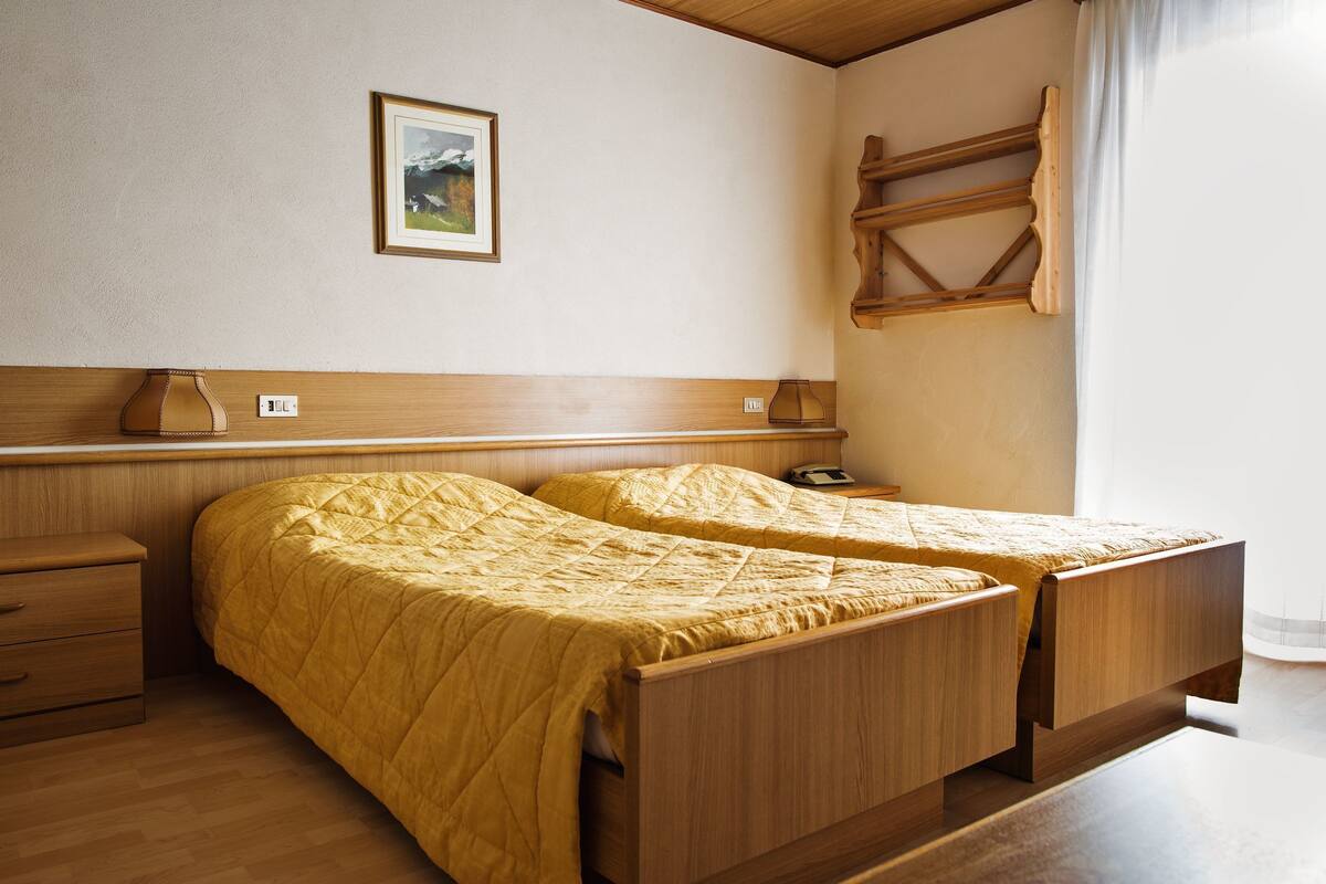 A room with two wooden beds, a bedside table, a lamp, a shelf, and a painting on the wall.