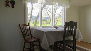 Un coin repas avec une table dressée pour deux personnes, une fenêtre donnant sur des arbres et des collines, et une décoration murale.
