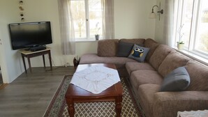 Un salon avec un canapé, un téléviseur posé sur un meuble, une petite table recouverte d’une nappe et une fenêtre avec des rideaux.