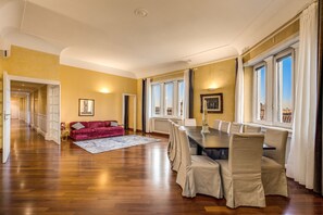 Una habitación amplia con suelo de madera, una mesa de comedor con sillas, un sofá rojo y ventanas grandes con cortinas.