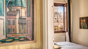 A bedroom with a bed, a radiator, a window with curtains, and a painting of a building.