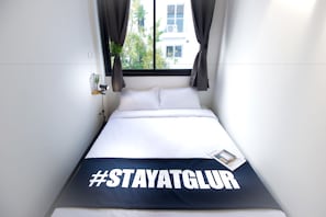 A bed with a #STAYATGLUR blanket, a small plant on the nightstand, and a window with curtains.
