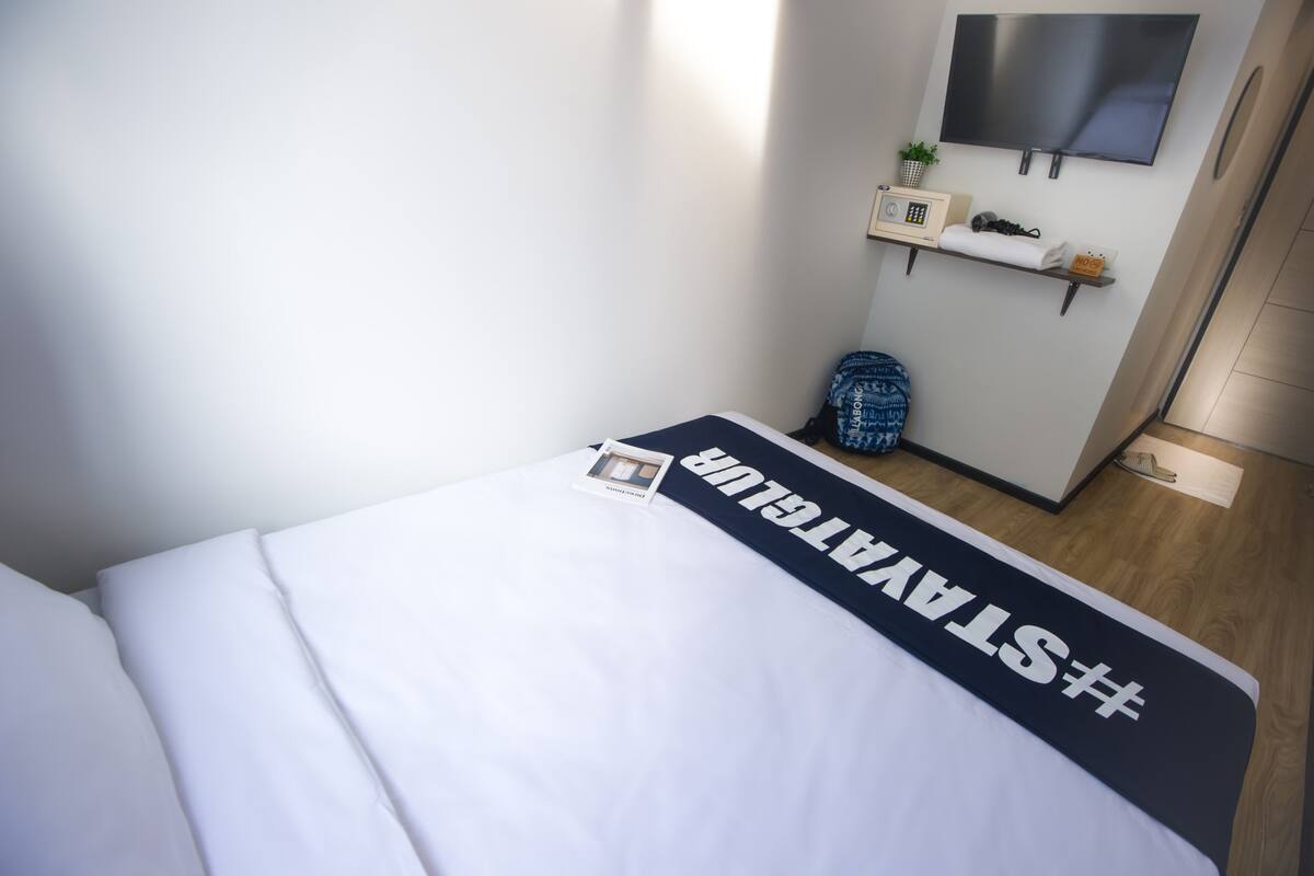 Una habitación de hotel con una cama, un televisor montado en la pared, una estantería con objetos y suelo de madera.