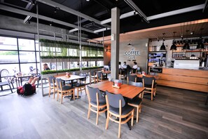 Ein modernes Café mit Holztischen und -stühlen, einer Theke mit einer Kaffeemaschine und großen Fenstern mit viel Grün.