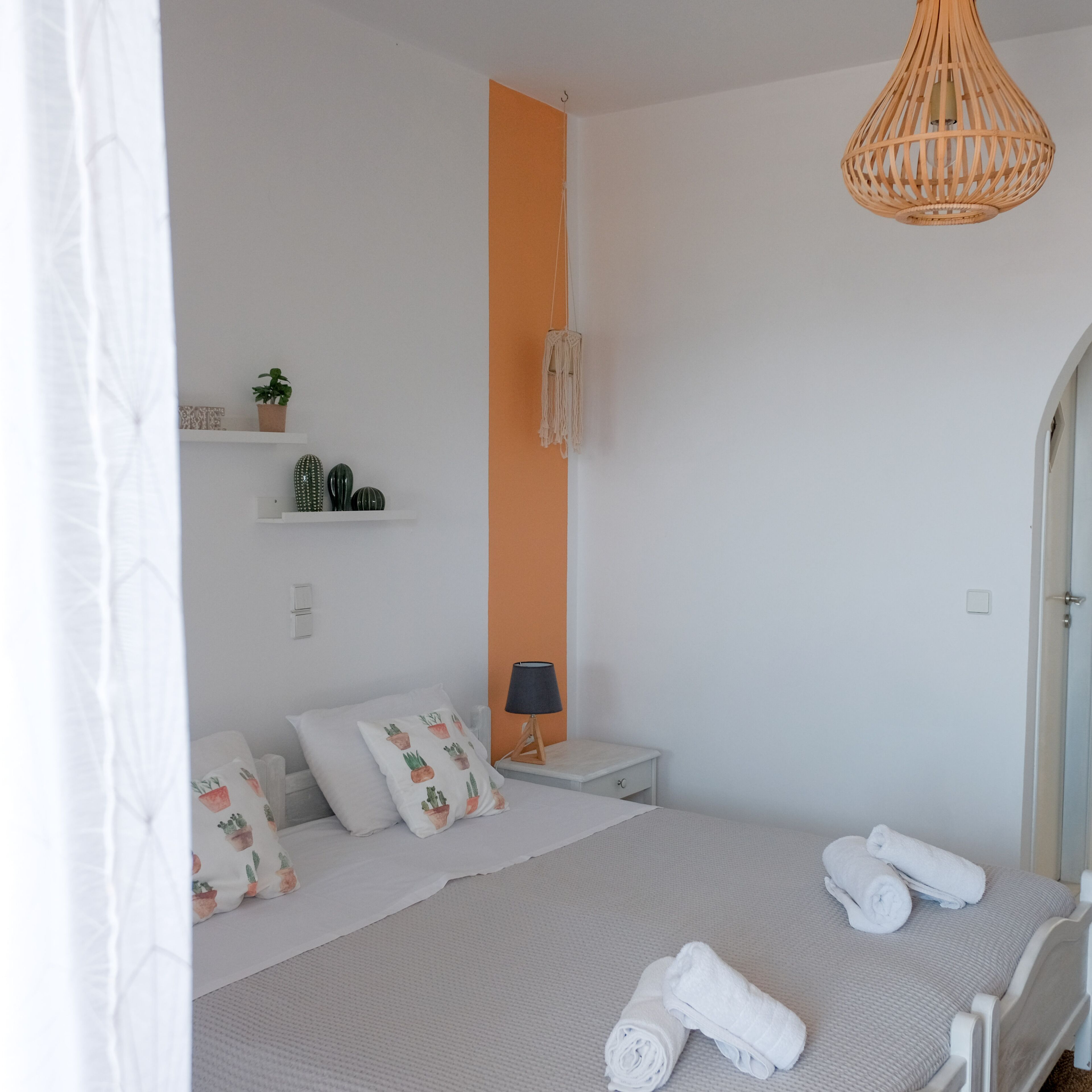 A bedroom with a bed, a bedside table, a lamp, a wall-mounted shelf with decorative items, and a hanging light fixture.