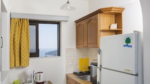 A compact kitchen with wooden cabinets, a white refrigerator, a coffee maker, and a view through the window. - Pefkides Aegina Apartments (Aegina)