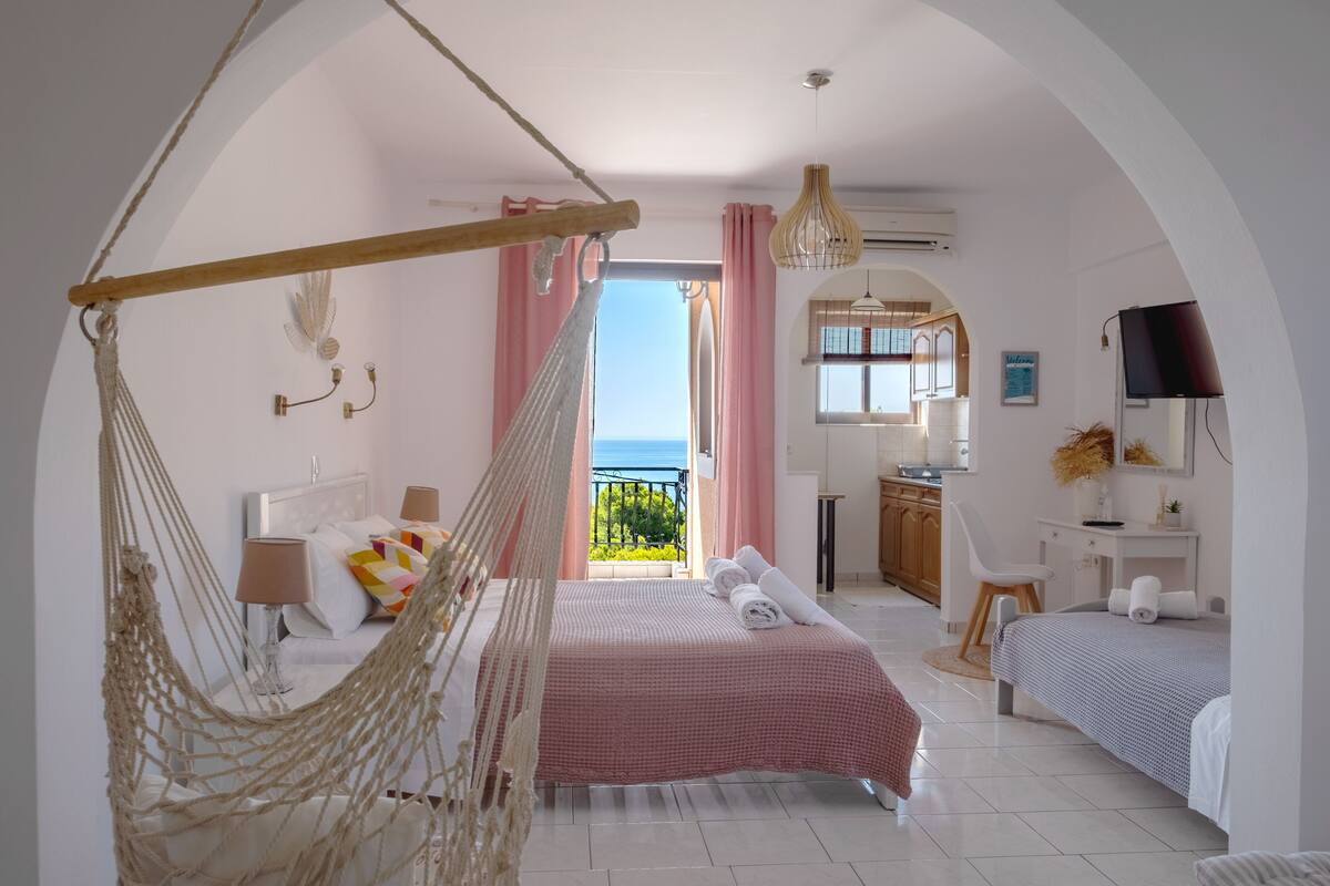 Un dormitorio con una hamaca, cama con ropa de cama rosa, un escritorio con silla y vista al mar.